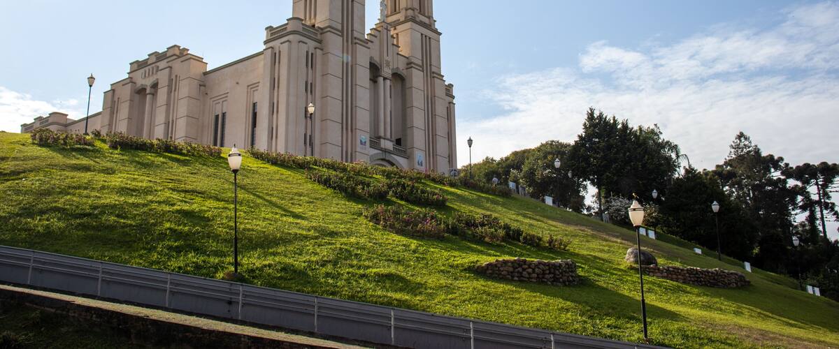 A large church with two towers and a cross on top, surrounded by a green hill and trees.