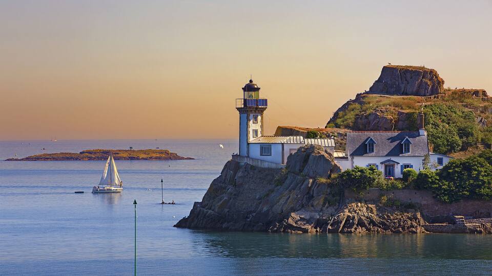 A lighthouse on a rocky cliff with a sailboat in the water and a distant island.