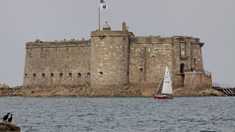 A large stone castle by the sea with a sailboat nearby.