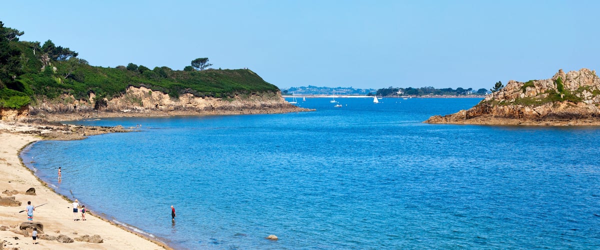 A coastal scene with a sandy beach, clear blue water, rocky outcrops, and distant sailboats.