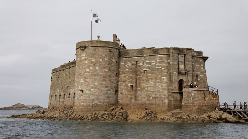 A large stone castle by the sea with a flag on top.