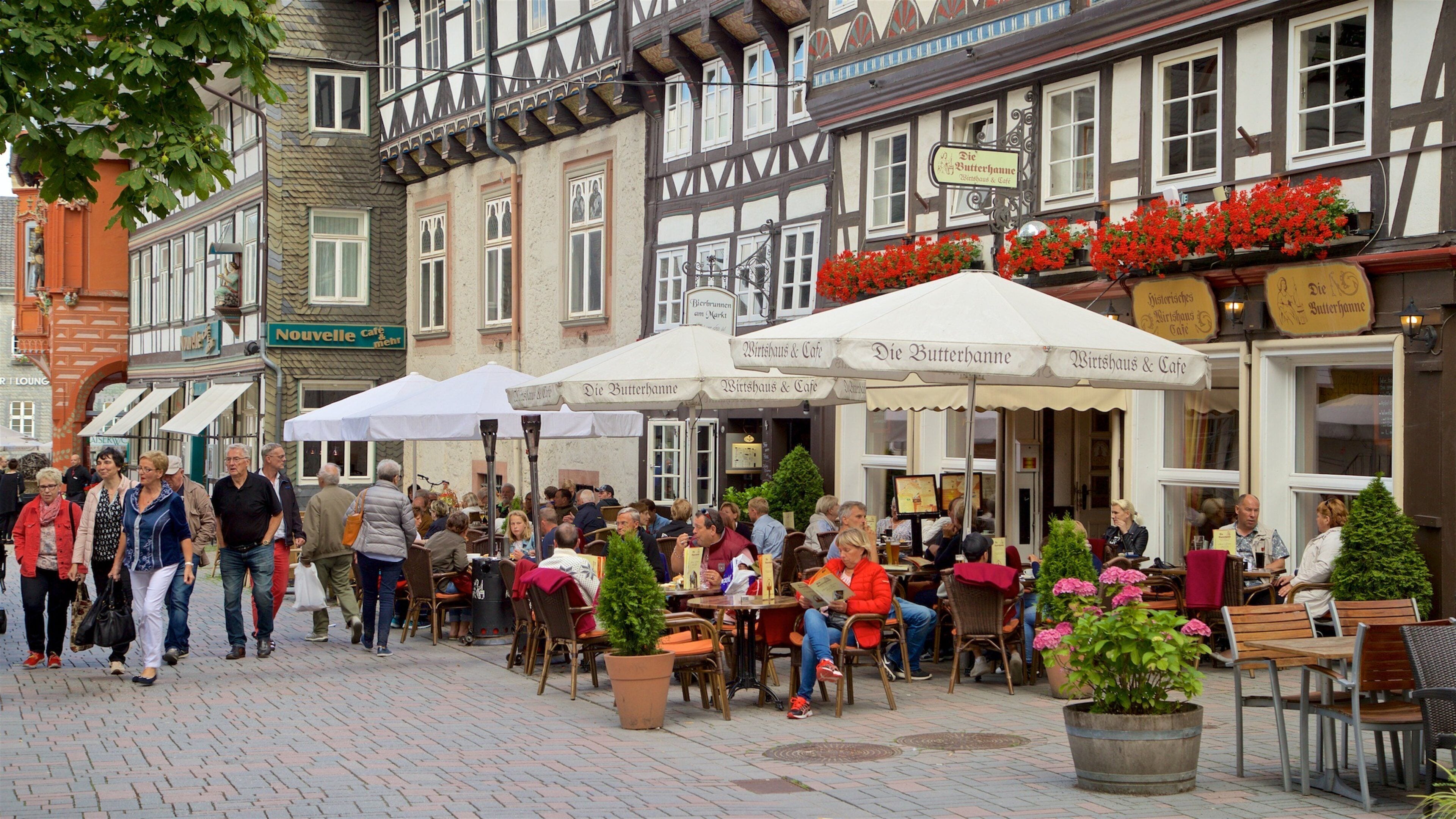 A bustling outdoor café area with traditional half-timbered buildings, outdoor seating, and red flowers.