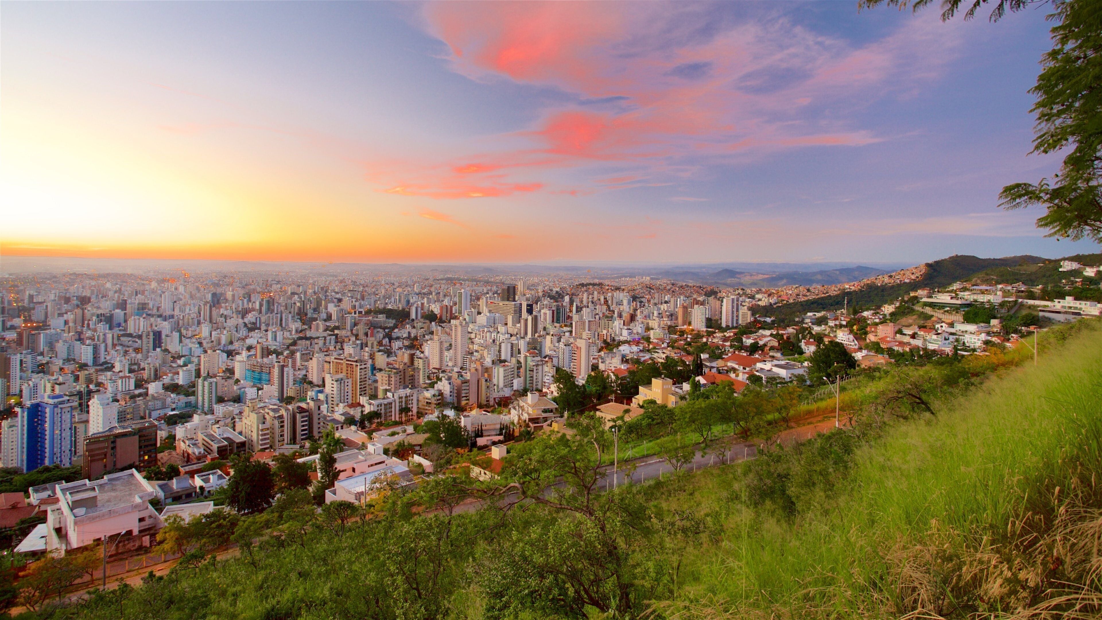 A cityscape at sunset with a dense urban area and a hillside with greenery.