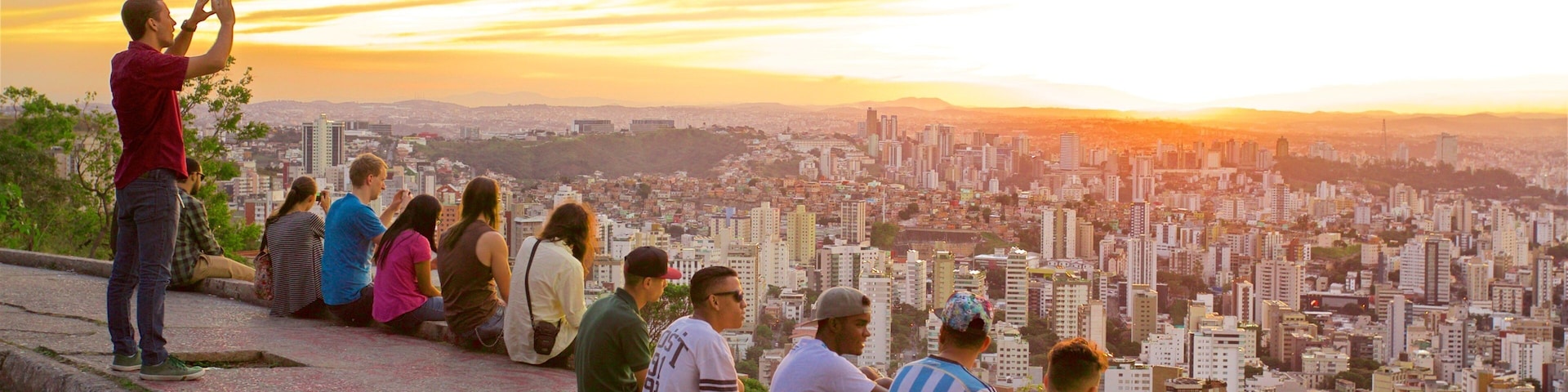Un groupe de personnes est assis sur une corniche de pierre, d’où elles contemplent un paysage urbain au coucher du soleil.