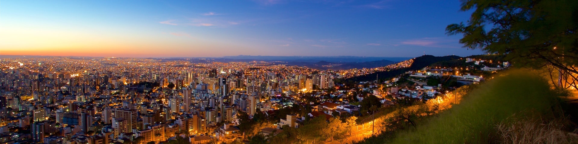 Vista urbana ao anoitecer, com luzes da cidade e uma colina em primeiro plano.