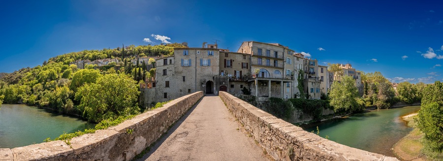 Un puente de piedra histórico que conduce a un edificio fortificado con múltiples ventanas y balcones, rodeado de exuberante vegetación y un río.