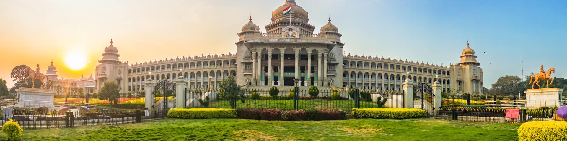 Panoramic view of Vidhana Soudha, the iconic government building in Bengaluru, India, captured during golden hour. The lush gardens and clear sky frame the historic architecture beautifully