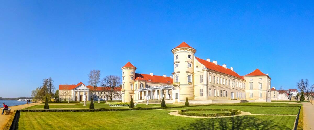A large historic building with a red roof and a tower by a lake.