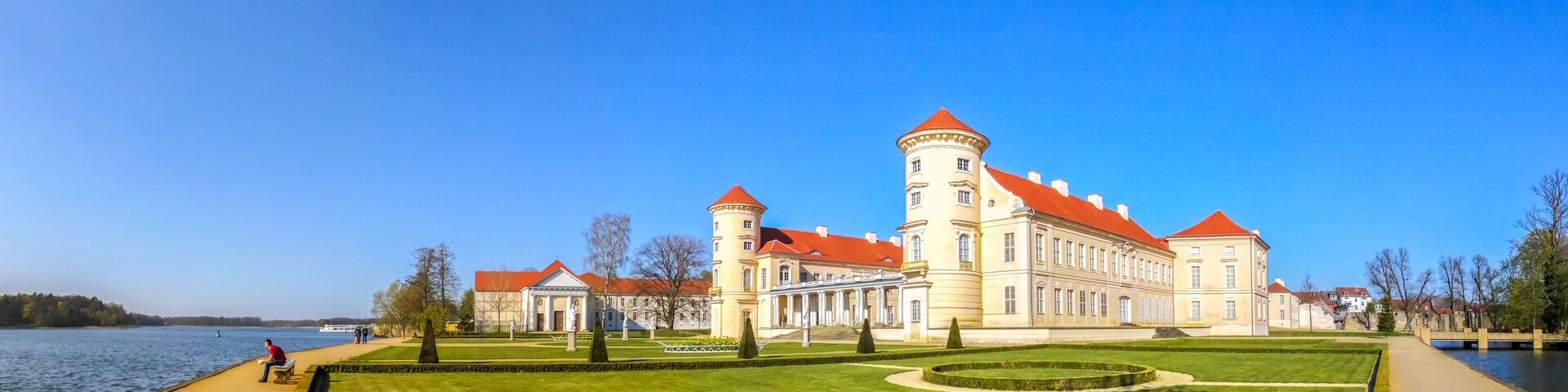 A large historic building with a red roof and a tower by a lake.