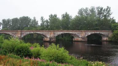 A stone bridge over a river with greenery and trees in the background.
