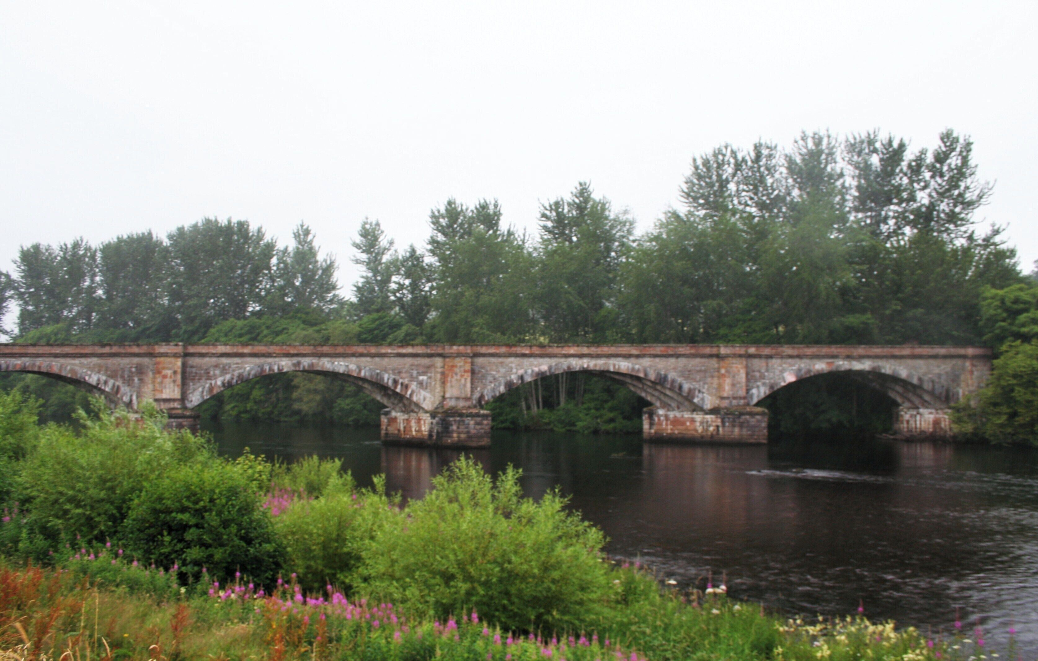 A stone bridge over a river with greenery and trees in the background.