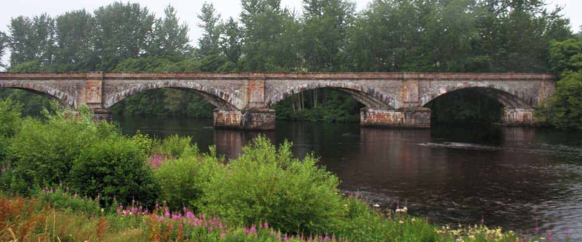 A stone bridge over a river with greenery and trees in the background.