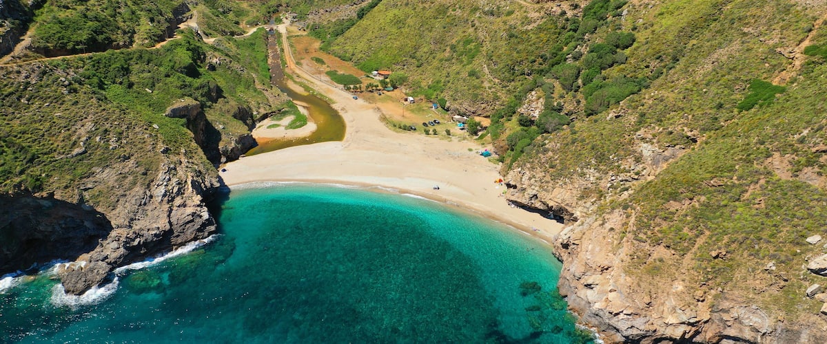 An aerial view of a coastal area with a sandy beach, clear turquoise waters, and surrounding green hills.