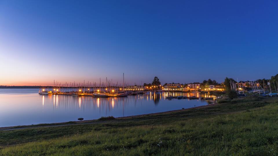 A calm lake at dusk with illuminated buildings and boats along the shore.