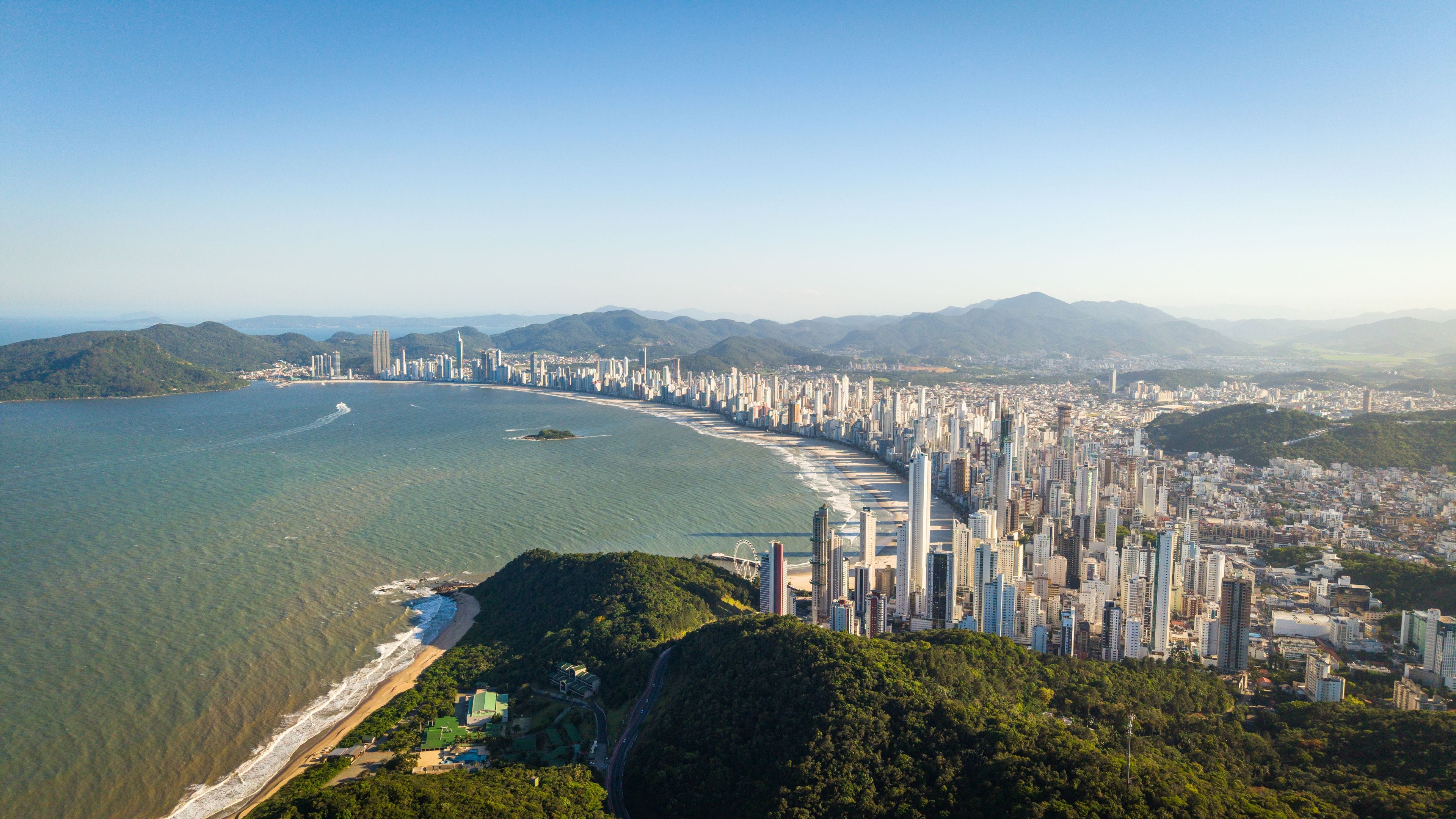 A coastal cityscape with a dense cluster of high-rise buildings along the beachfront, a clear blue sky, and surrounding hills.