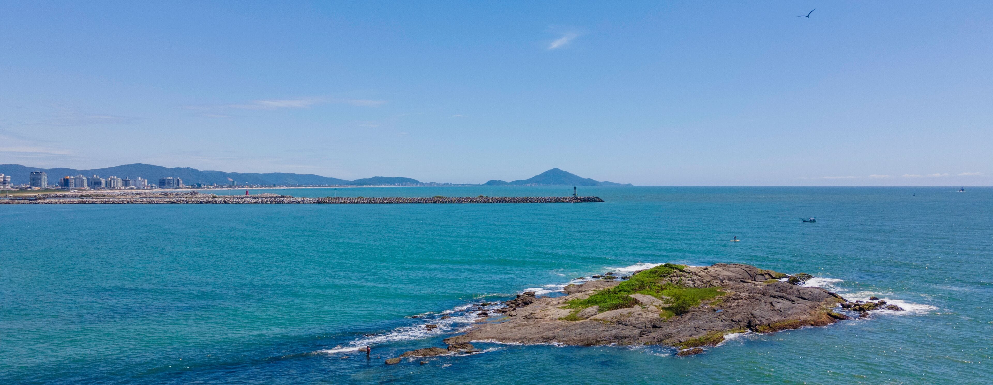 A coastal cityscape with a prominent rocky island, clear blue waters, and distant mountains.
