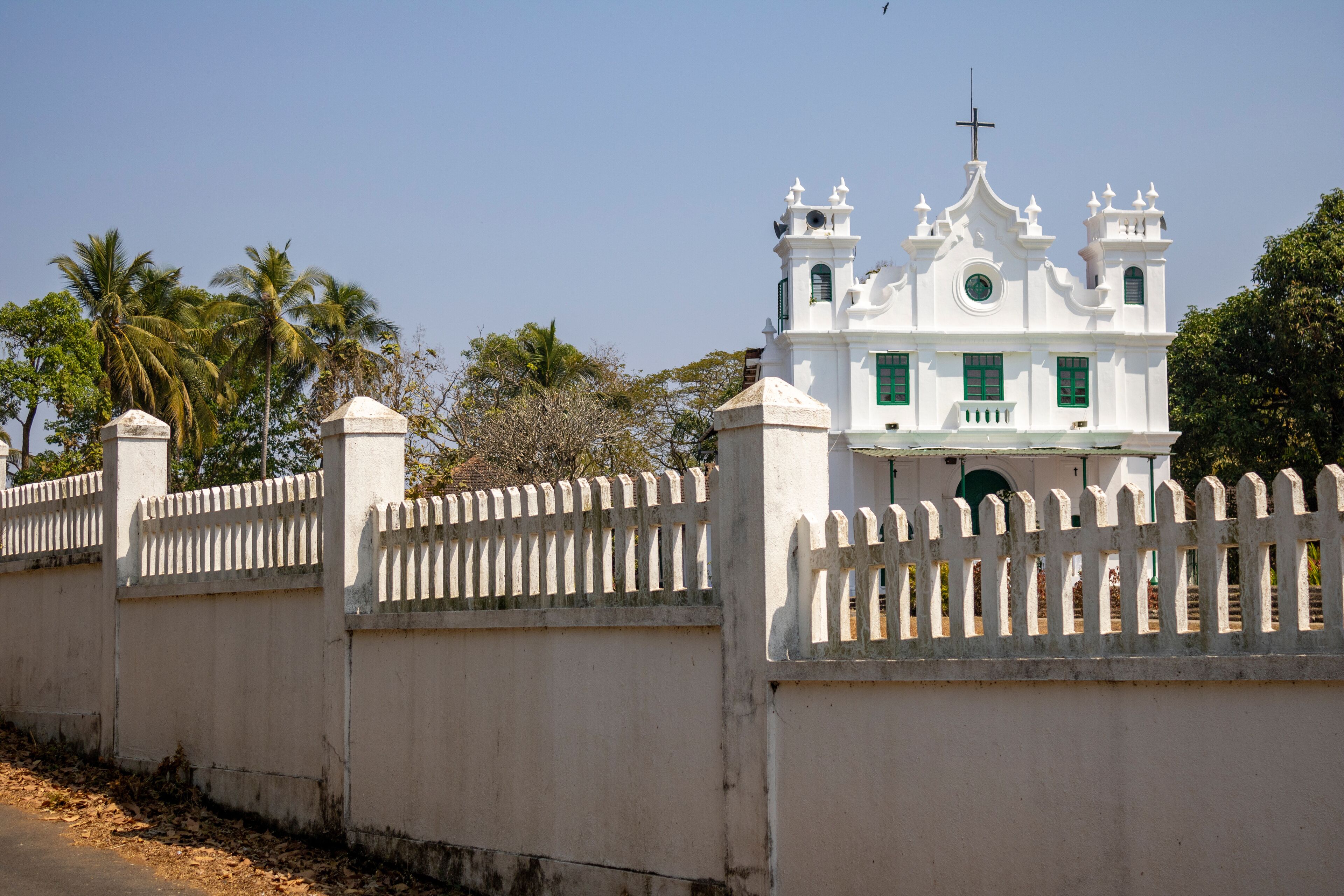 A white church with a cross on top, surrounded by a white picket fence and palm trees.