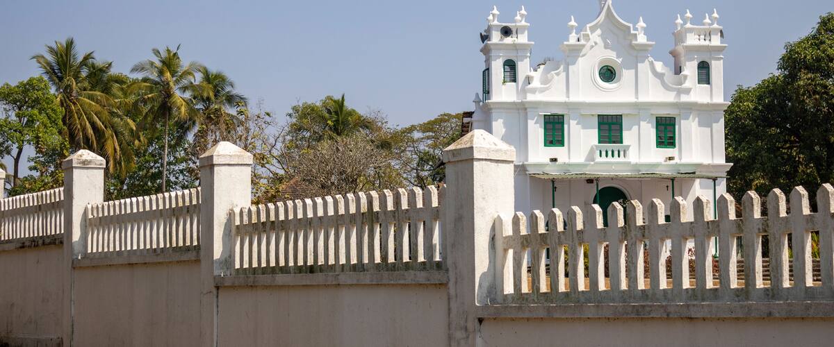 A white church with a cross on top, surrounded by a white picket fence and palm trees.