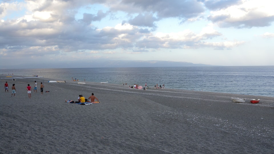 A beach with people sunbathing and swimming, boats on the water, and a cloudy sky.