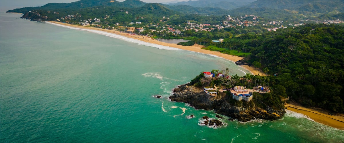 A coastal landscape with a beach, a small island with buildings, and a mountainous backdrop.