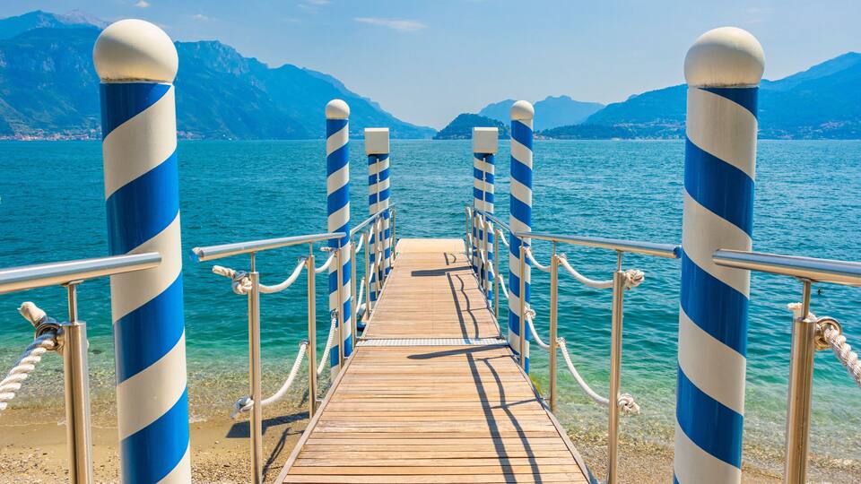 A wooden pier with blue and white striped bollards leading to a calm body of water, surrounded by mountains.