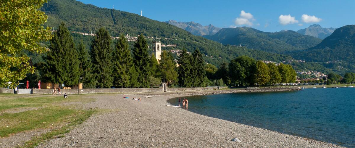 A pebble beach with a church tower, trees, and mountains in the background.