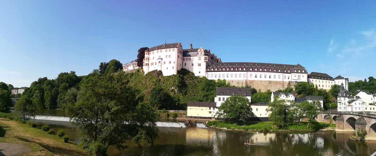 A castle on a hill overlooking a river, surrounded by lush greenery and historic buildings.