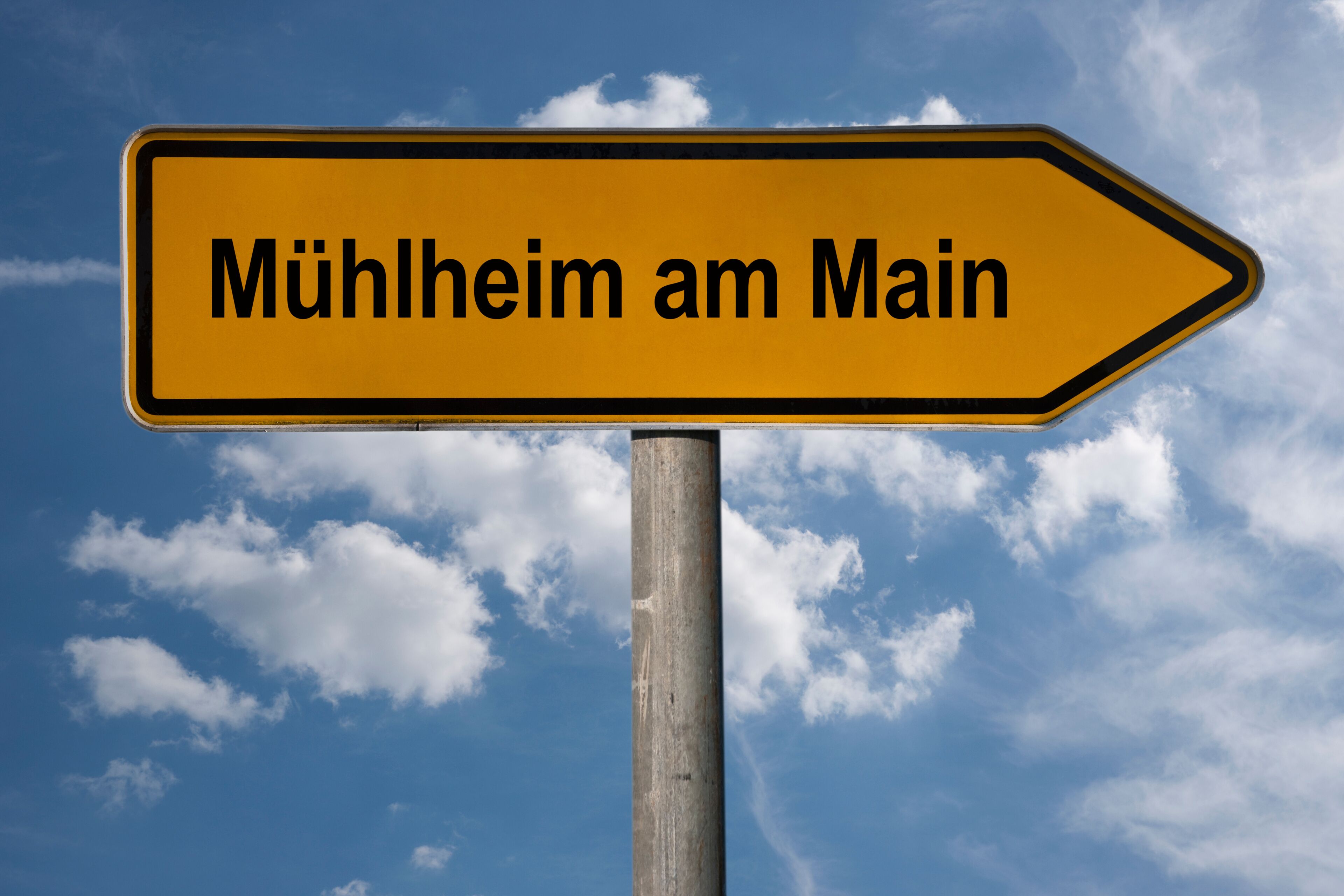 A directional sign with the text "Mühlheim am Main" against a blue sky with clouds.