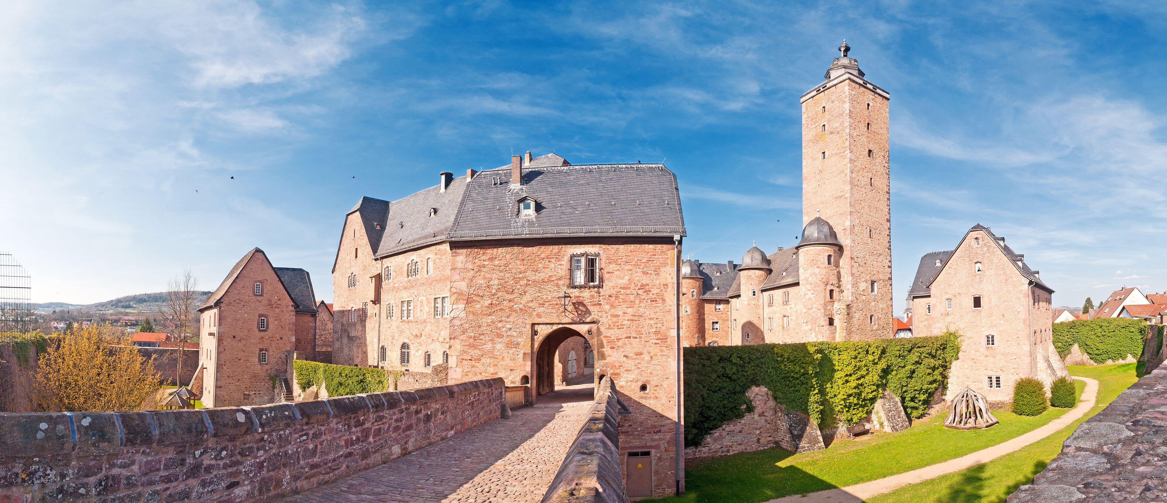 A historic castle with a prominent tower, a paved entrance, and surrounding greenery.