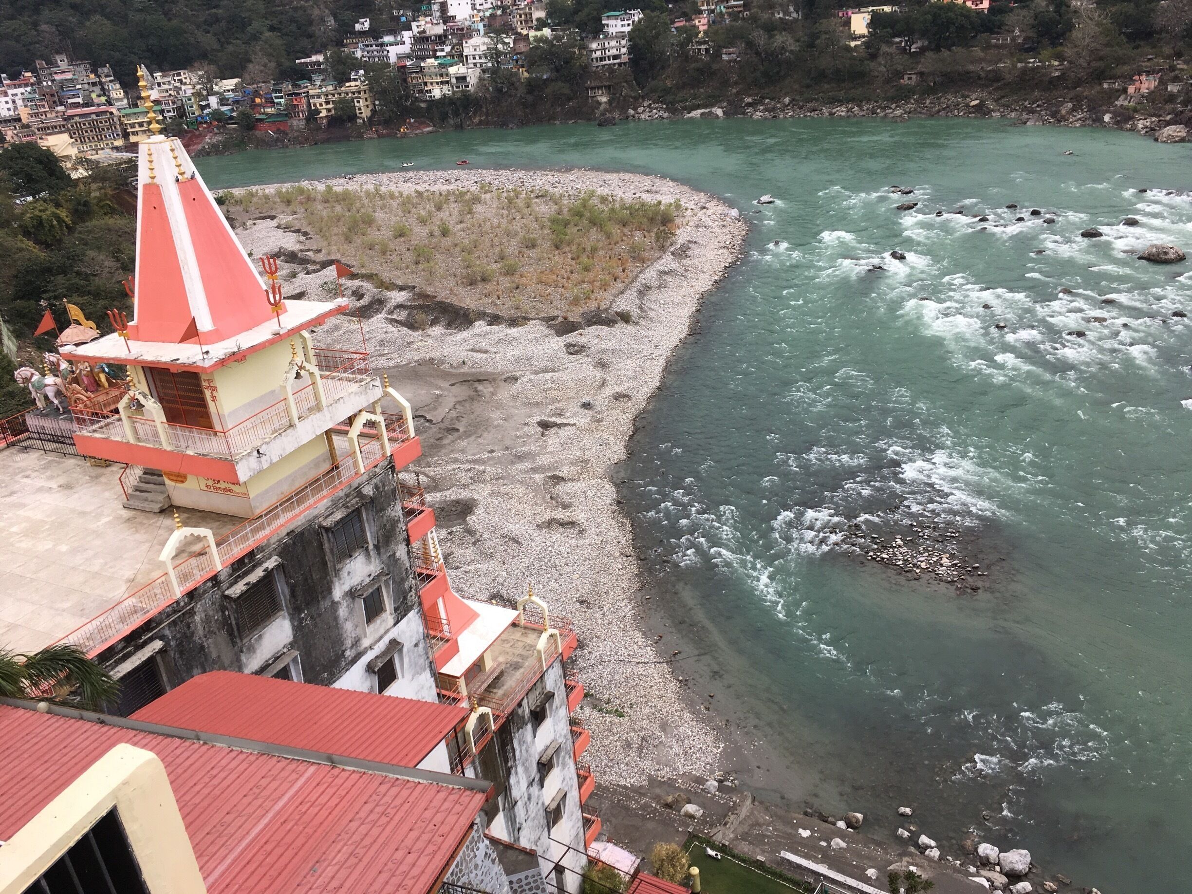 A river with a rocky shoreline, a temple with a red roof, and a hillside with buildings.