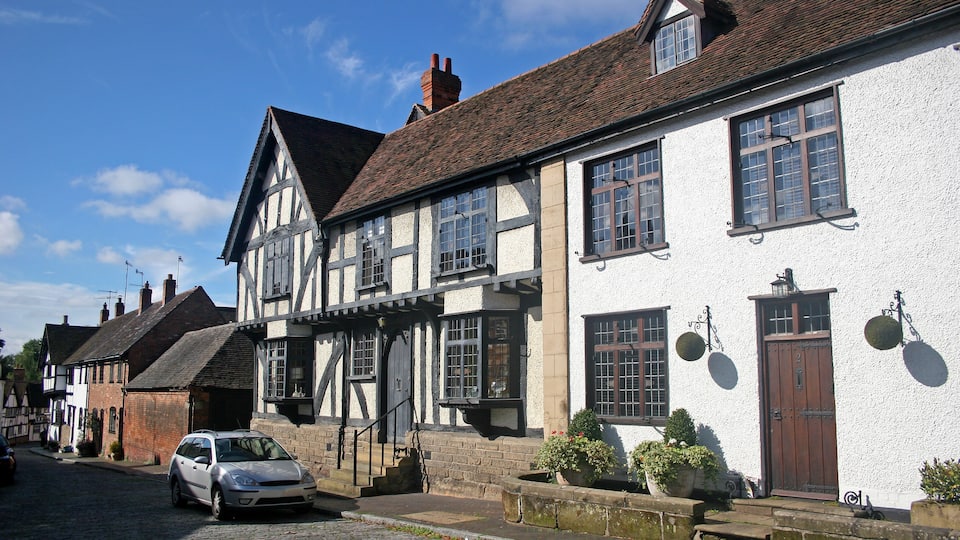 A cobblestone street lined with traditional half-timbered houses, a parked car, and a clear blue sky.