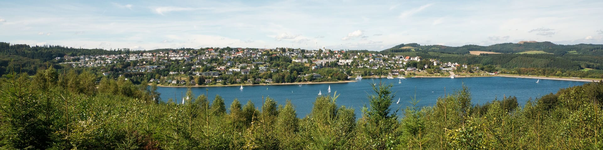 Wide view over Lake Sorpesee with sailboats and the dam with Langscheid in the background in front of a cloudy sky in Sundern, Sauerland, Germany