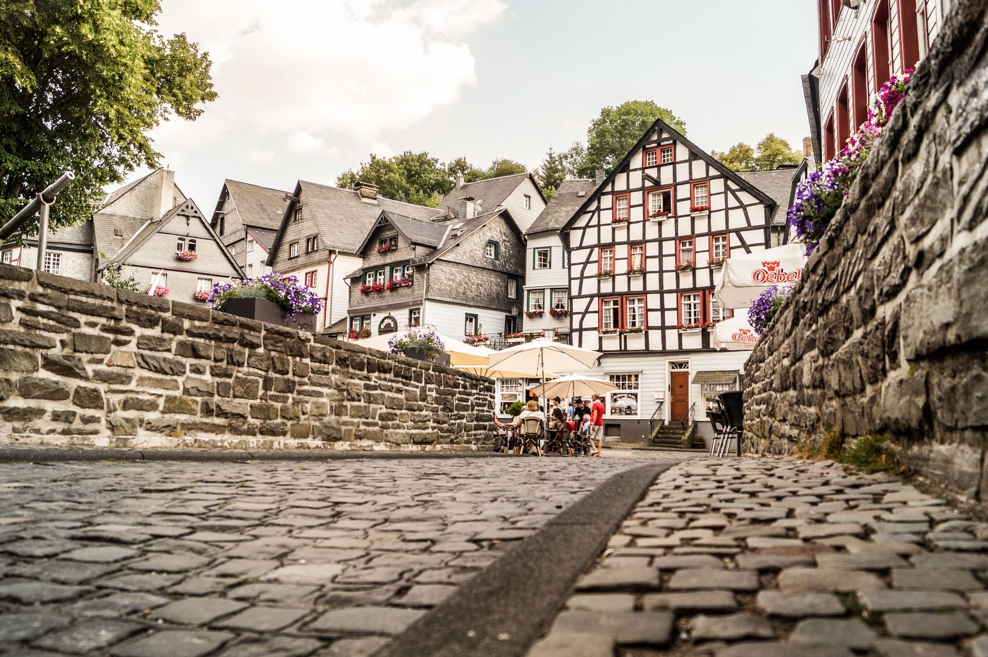 Una calle adoquinada en un pueblo histórico con casas tradicionales de entramado de madera, un muro de piedra y un café con sombrillas.