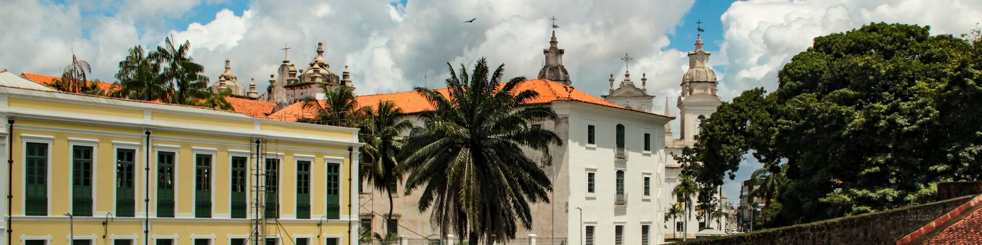 A historic building with a yellow facade and green shutters, a stone wall, and palm trees under a partly cloudy sky.