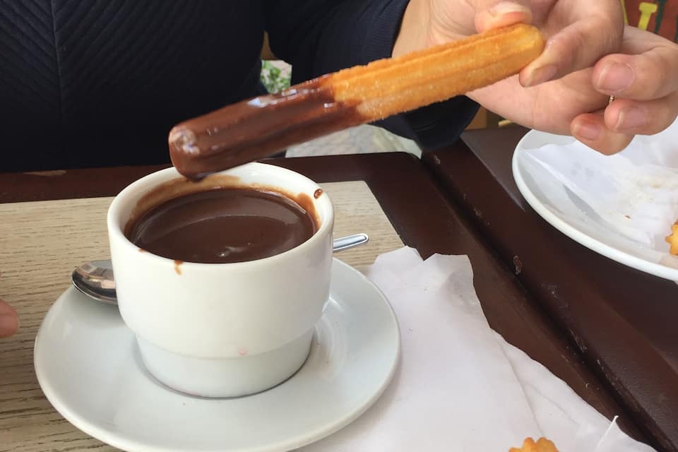 A person holding a churro dipped in chocolate, a white cup of hot chocolate, and a plate with churros.