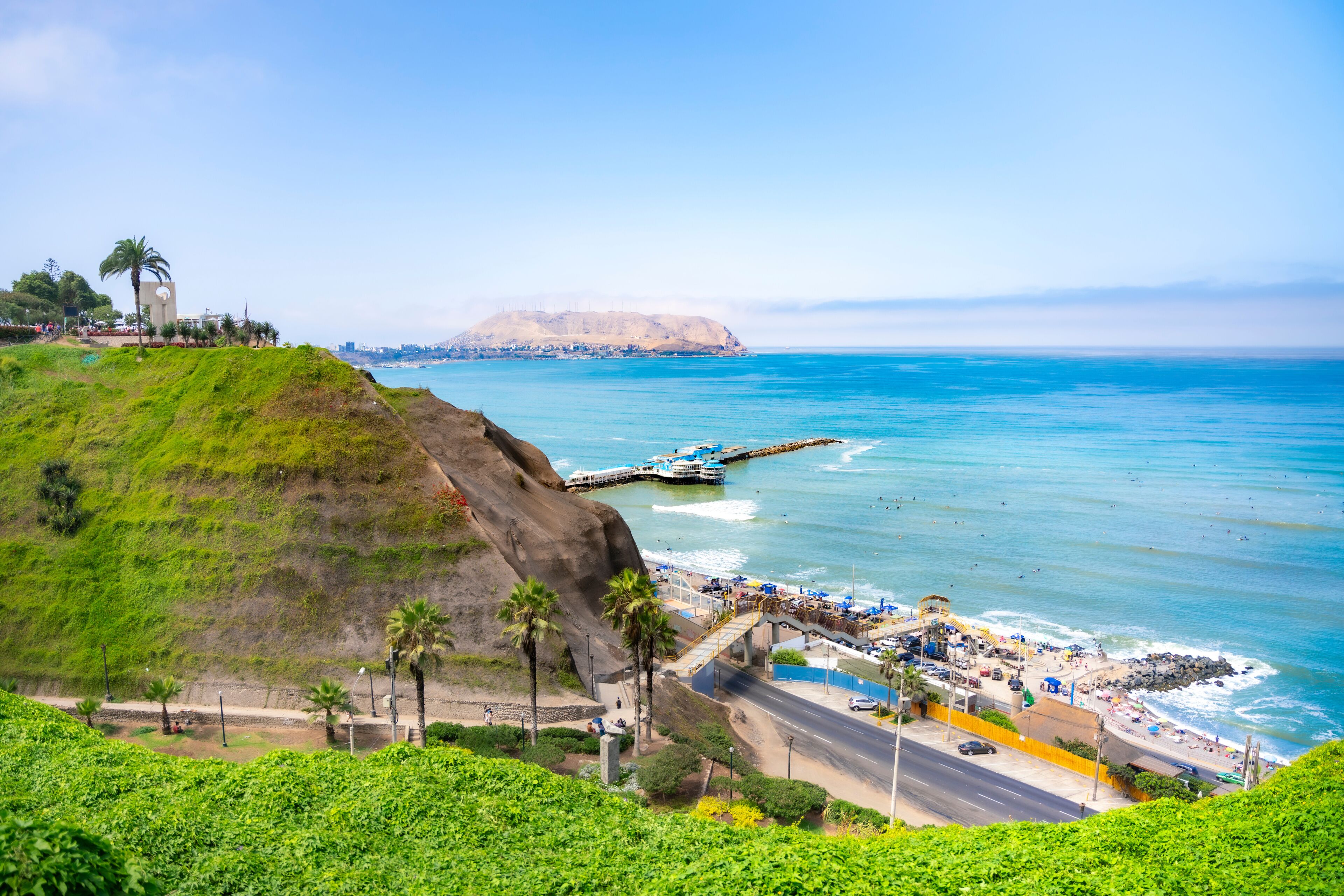 A coastal cityscape with a prominent hill, a pier extending into the sea, and a distant mountain.