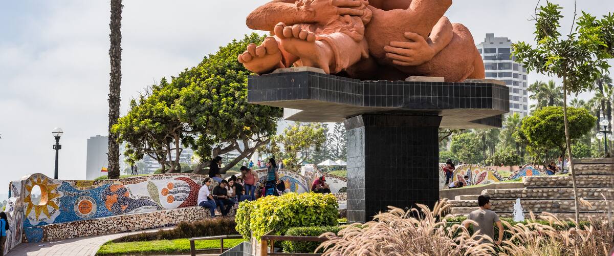 Una gran escultura al aire libre de dos figuras abrazándose, ubicada sobre una plataforma de baldosas en un parque público con palmeras y un muro de mosaicos al fondo.