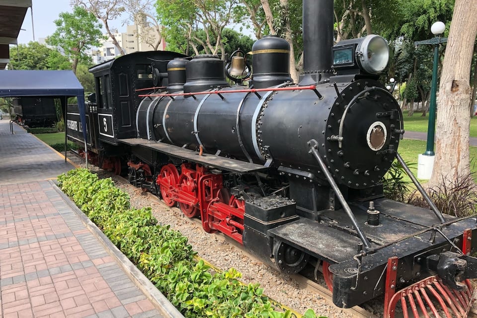 Uma locomotiva a vapor antiga, com rodas vermelhas e corpo preto, estacionada sobre um trilho de tijolos, com vegetação de ambos os lados.