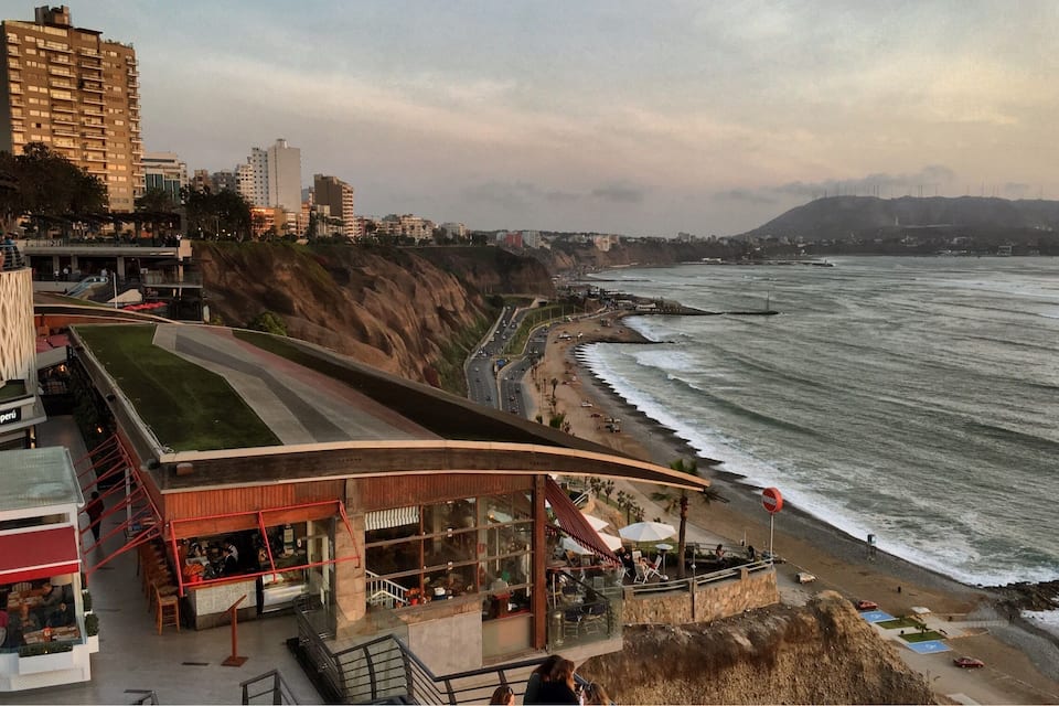 A coastal cityscape with a restaurant overlooking the beach, a cliffside, and a view of the ocean.
