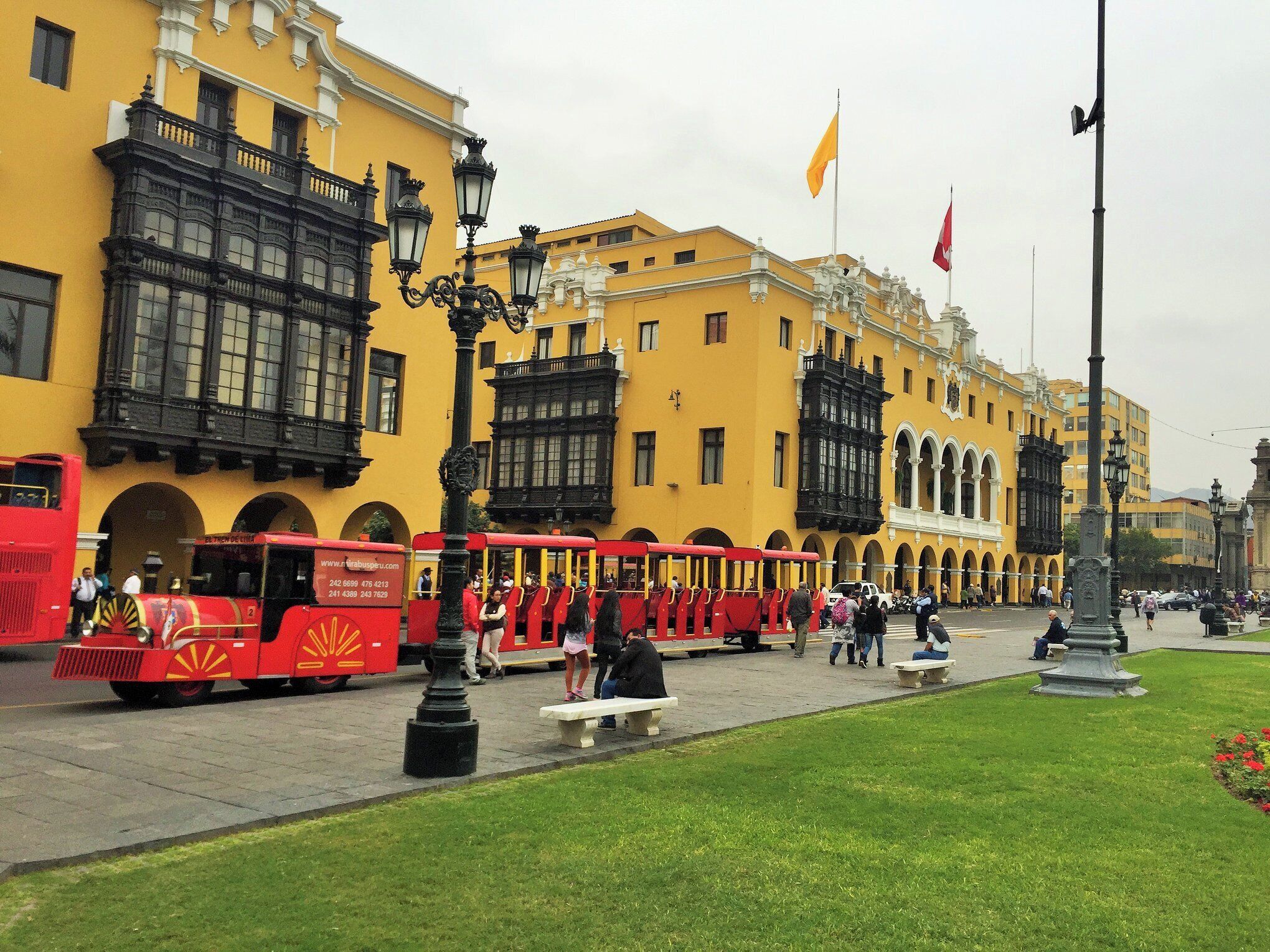 Un tranvía rojo con pasajeros, edificios amarillos y una estatua en una plaza.