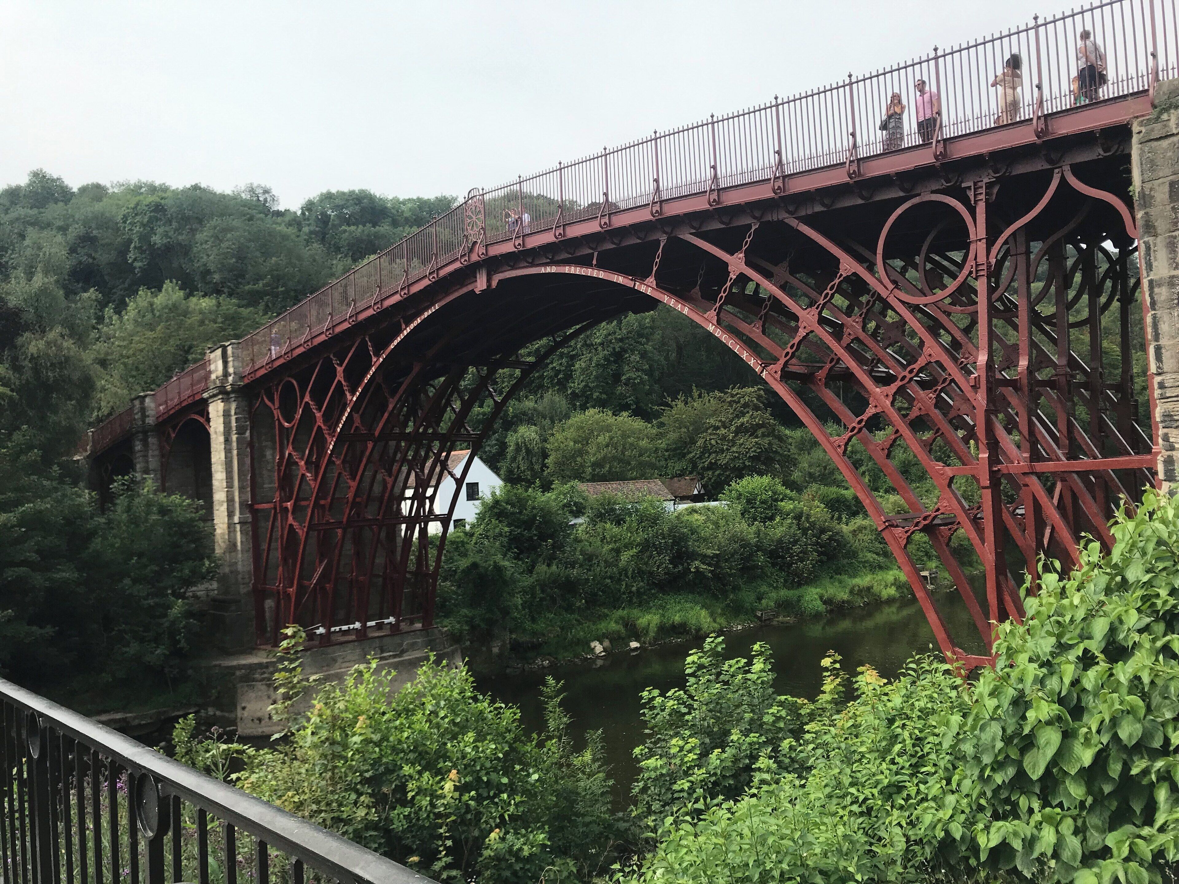 A red metal bridge with ornate designs spanning over a river, surrounded by lush greenery.