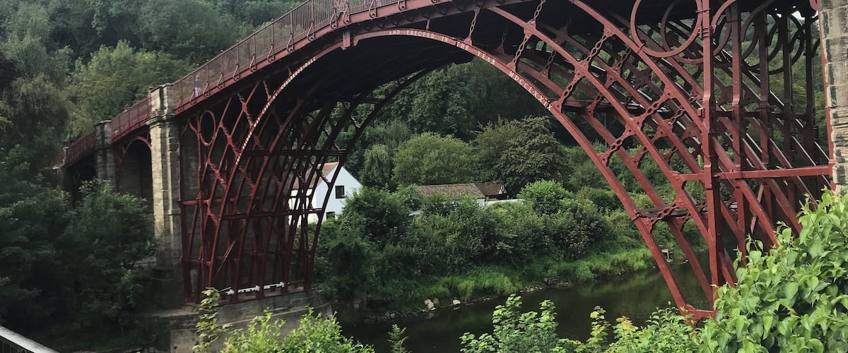A red metal bridge with ornate designs spanning over a river, surrounded by lush greenery.