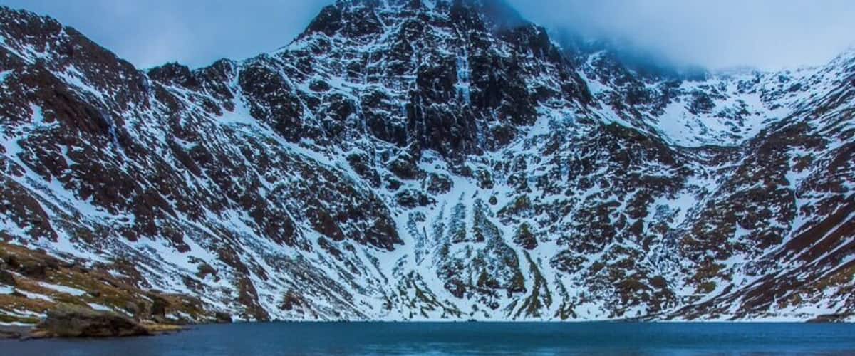 A mountain range with snow-covered peaks and a clear lake with rocks in the foreground.