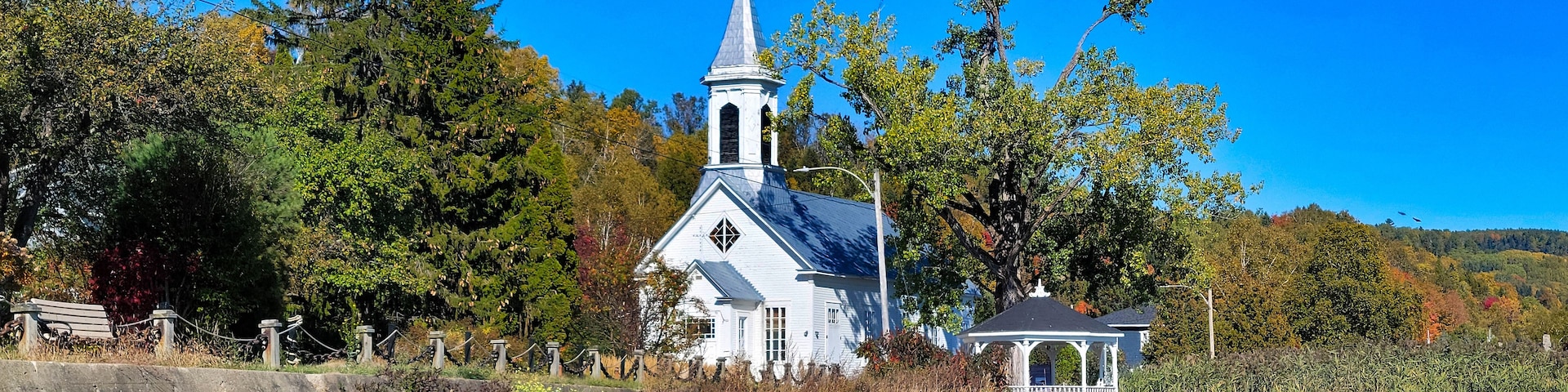 A white church with a steeple, surrounded by trees and a small gazebo.