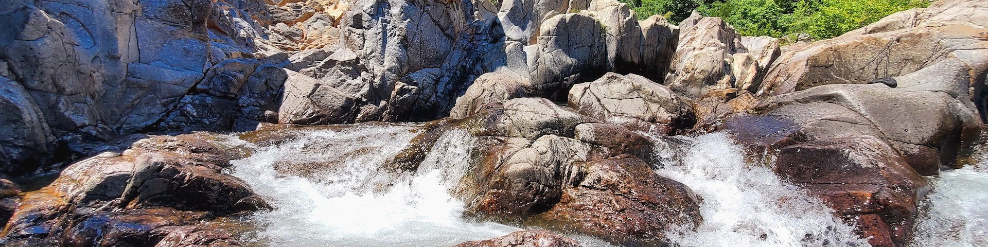 Un lecho fluvial pedregoso con aguas cristalinas, rodeado de grandes rocas y abundante vegetación.