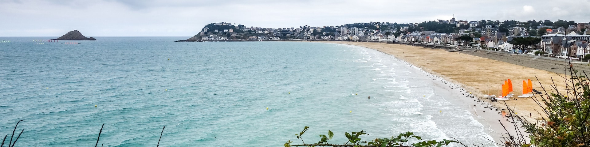Un paisaje costero con una playa arenosa, olas del océano y una colina distante con edificios.