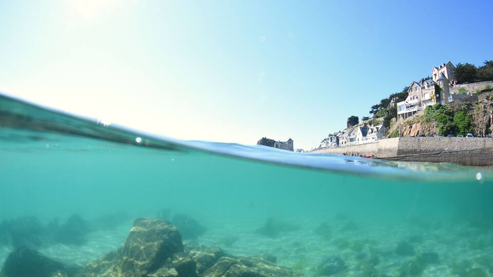 A coastal scene with clear turquoise water, rocky seabed, and a row of houses on a hillside.
