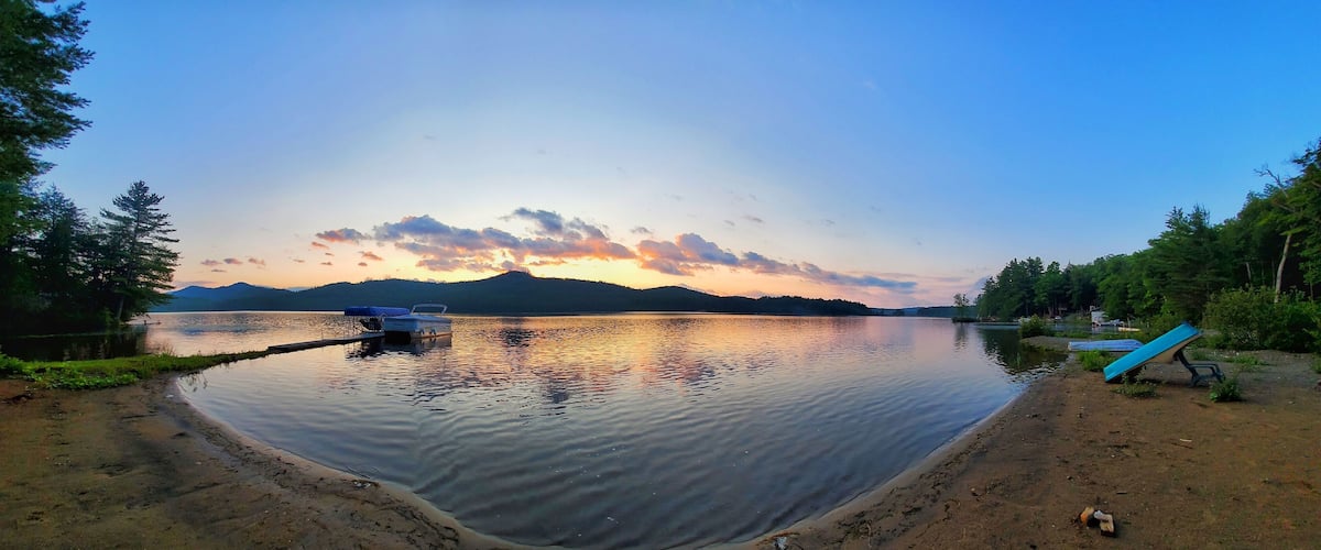 A calm lake at sunset with boats docked, surrounded by trees and mountains.