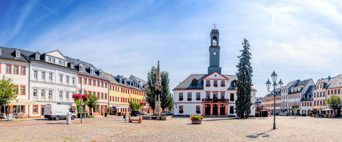 A cobblestone square with historic buildings, a church with a tall steeple, and a statue in the center.