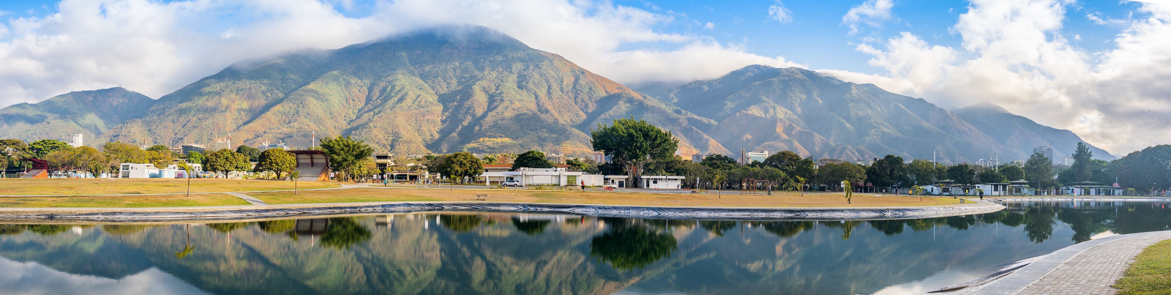 A calm lake reflecting the surrounding landscape, with a mountain range in the background and a park with buildings and trees in the foreground.